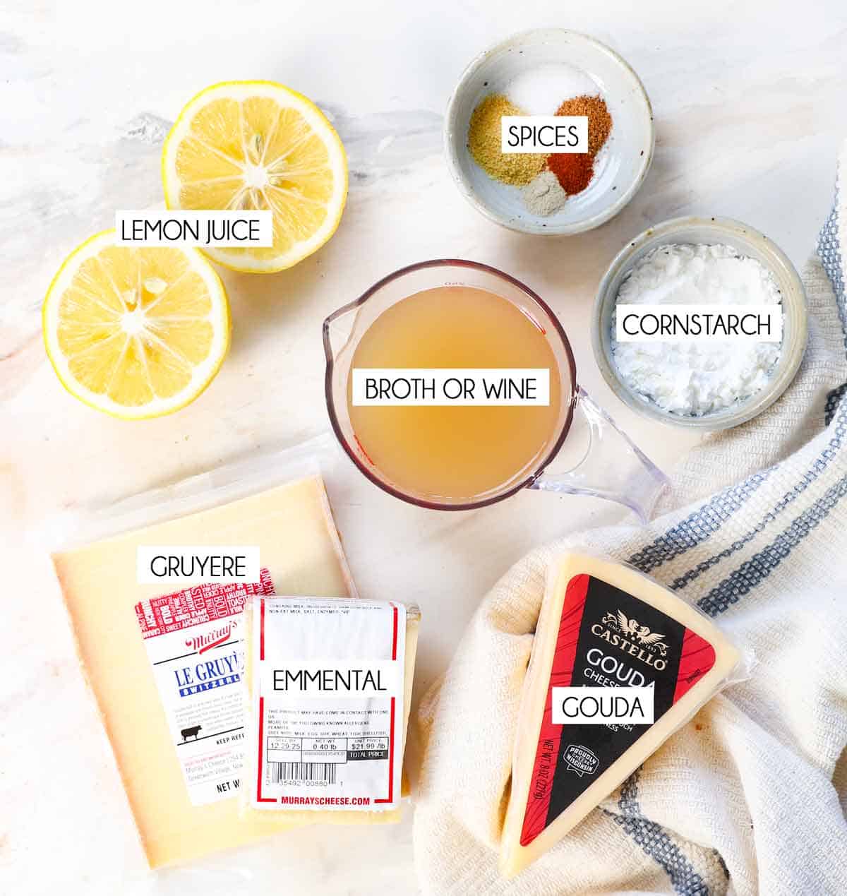 top view of fondue ingredients: Gruyere cheese, Emmental cheese, gouda, cornstarch, nutmeg, paprika, salt, ground mustard, and lemon juice