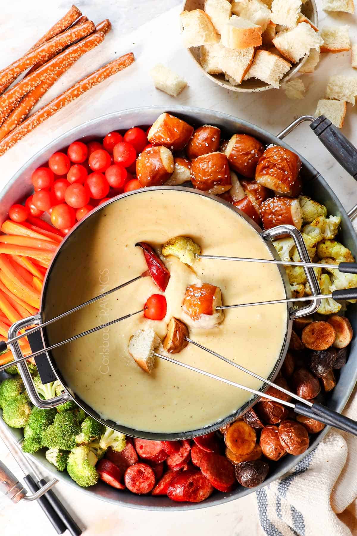top view of cheese fondue with a tray of dippers: bread, pretzels potatoes, broccoli, cauliflower, carrots, sausage, tomatoes