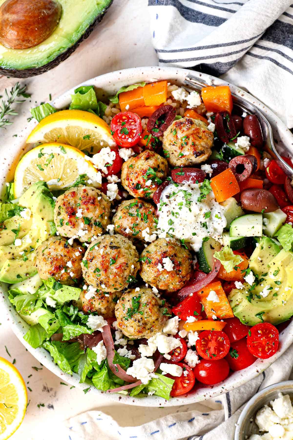 top view of baked chicken meatballs being served Greek style in a bowl with rice, Kalamata olives, tomatoes, feta, avocados, lemon and tzatziki