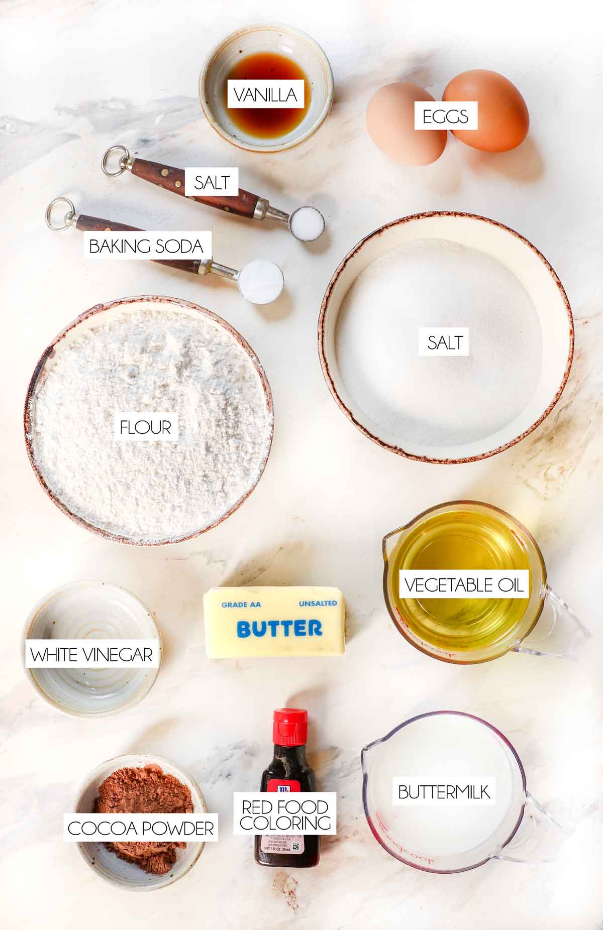 top view of red velvet cake ingredients: flour, cocoa powder, butter, sugar, vegetable oil, buttermilk, red food coloring, baking soday, salt, eggs, white vinegar