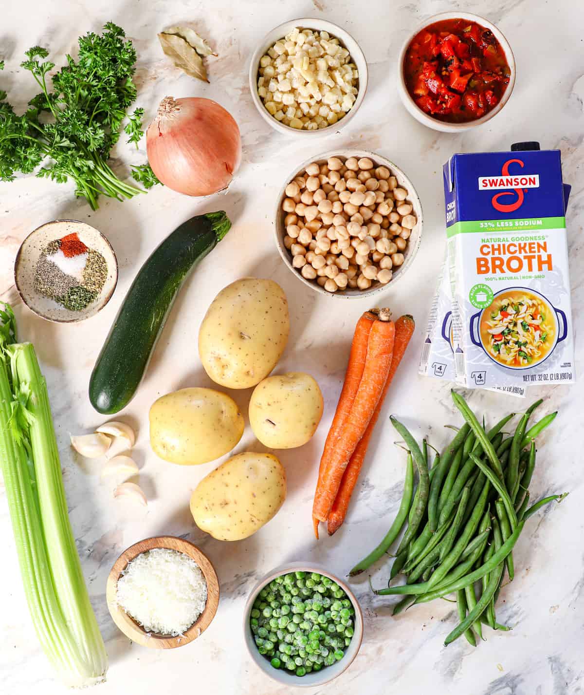 top view of vegetable soup ingredients: broth, chickpeas, potatoes, carrots, green beans, peas, corn, potatoes, zucchini, onions, celery, parsley