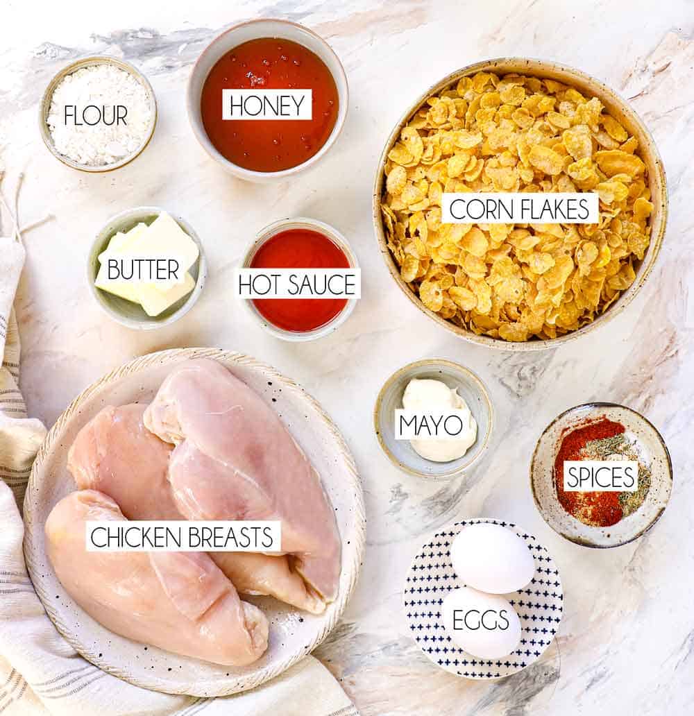 top view of hot honey chicken ingredients: chicken breasts, honey, corn flakes, hot sauce, eggs, flour, spices