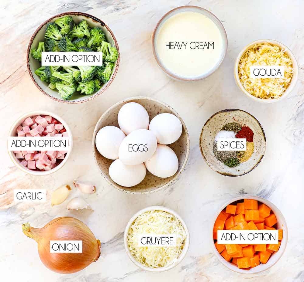 top view of crustless quiche ingredients: eggs, heavy cream, spices, Gruyere cheese, Gouda cheese, onion, garlic and optional add-ins: broccoli, ham, sweet potatoes