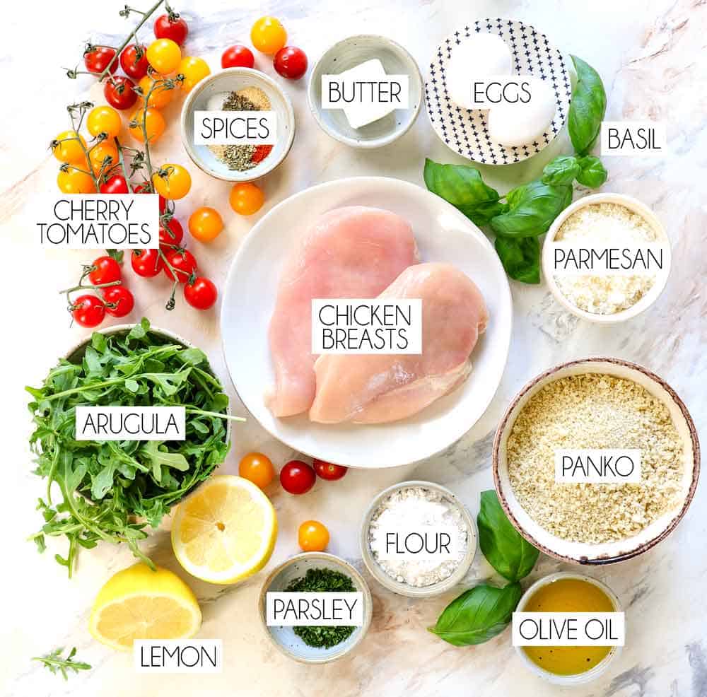 Top view of chicken Milanese recipe ingredients: chicken breasts, panko, flour, eggs, parsley, lemon zest, arugula, tomatoes, basil, olive oil