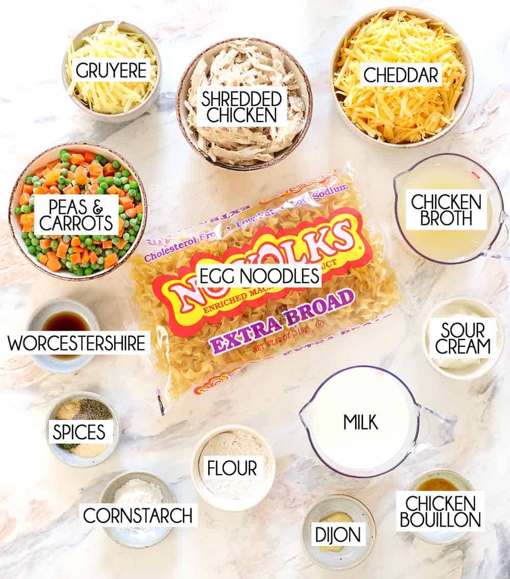 top view of chicken noodle casserole ingredients: egg noodles, shredded chicken, chicken broth, milk, flour, sour cream, Dijon, spices, peas and carrots, cheddar cheese, Gruyere cheese