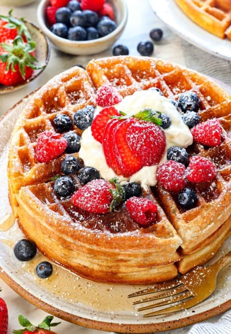 Belgian waffles stacked on a plate showing how fluffy they are, served with syrup, whipped cream, and berries