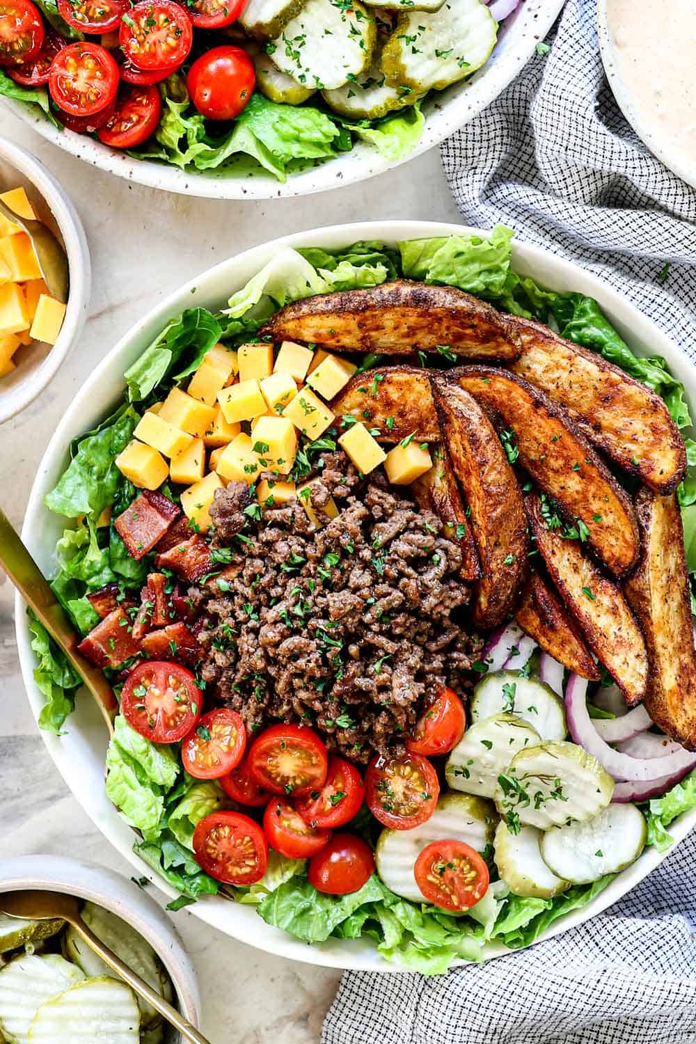 showing how to make burger bowls by assembling the bowls with lettuce on the bottom, then ground beef, tomatoes, pickles, red onion, cheese and potato wedges