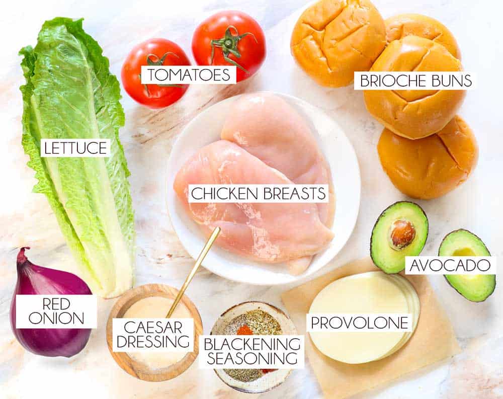 top view of chicken sandwich recipe ingredients: chicken breasts, brioche buns, provolone cheese, Romaine lettuce, tomato, red onion, avocado Caesar dressing, spices