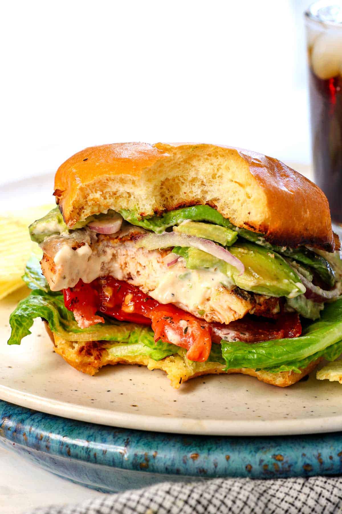 a bite taken out of spicy chicken sandwich showing the layers of lettuce, tomato, grilled chicken breast, avocado, red onion and bun