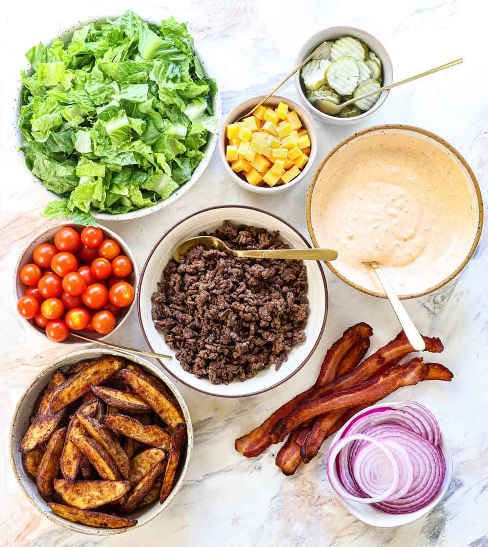 top view of burger bowl ingredients: lettuce, ground beef, spices, tomatoes, cheese, pickles, red onion, bacon, potato wedges