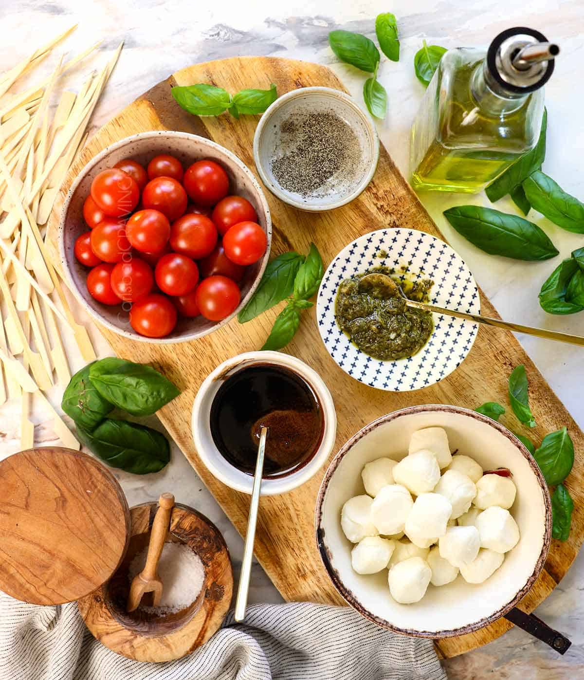 Top view of Caprese skewers recipe ingredients of cherry tomatoes, mozzarella balls, basil, pesto, olive oil, balsamic glaze, salt and pepper, and mini bamboo skewers