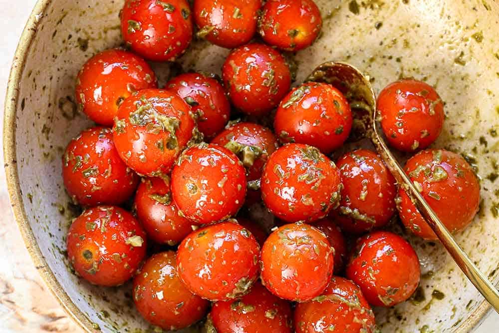 showing how to make Caprese skewers by tossing the cherry tomatoes in basil pesto in a bowl