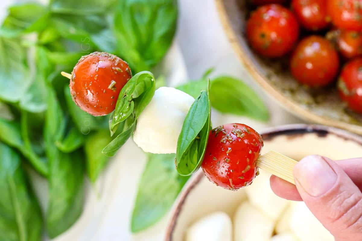 showing how to make Caprese skewers by threading the skewers with a cherry tomato, then basil, the mozzarella, then basil, then cherry tomato