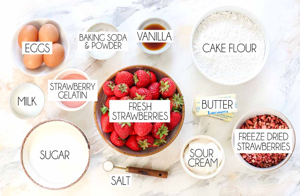 top view of strawberry cake ingredients: fresh strawberries, freeze dried strawberries, strawberry gelatin, cake flour, sugar butter, sour cream, vanilla, eggs, salt, baking powder, baking soda