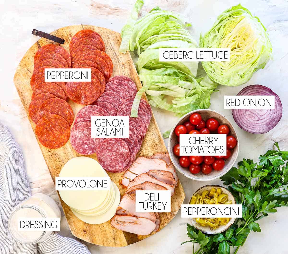 top view of Grinder Salad ingredients: iceberg lettuce, salami, pepperoni, deli turkey, provolone cheese, red onion, cherry tomatoes, pepperoncini