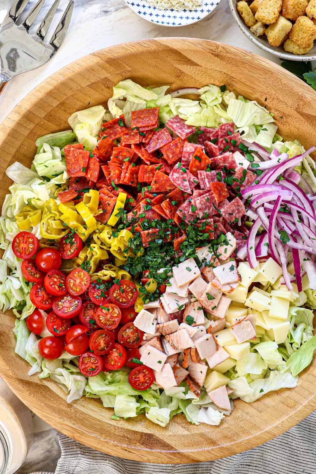 top view of Grinder salad ingredients in a bowl: iceberg lettuce, salami, pepperoni, deli turkey, red onions, tomatoes, pepperoncini, and Parmesan