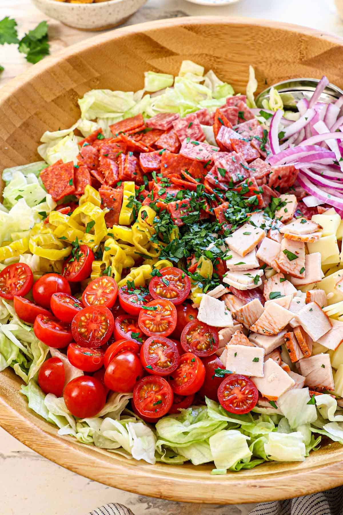 showing how to make Grinder salad by adding iceberg lettuce, salami, pepperoni, deli turkey, provolone cheese, cherry tomatoes, pepperoncini, red onions to a wood bowl