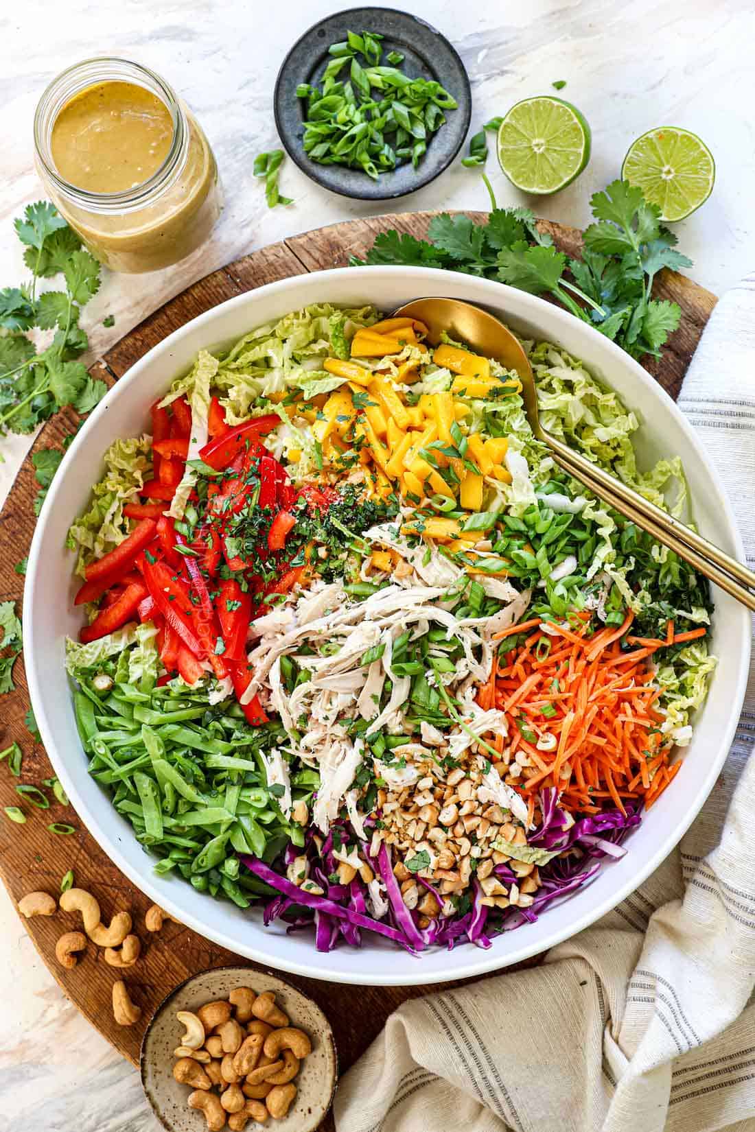 showing how to make Thai chicken salad recipe by adding the cabbages, chicken, bell peppers, carrots, snow peas, peanuts, mango, green onions and cilantro to a large bowl