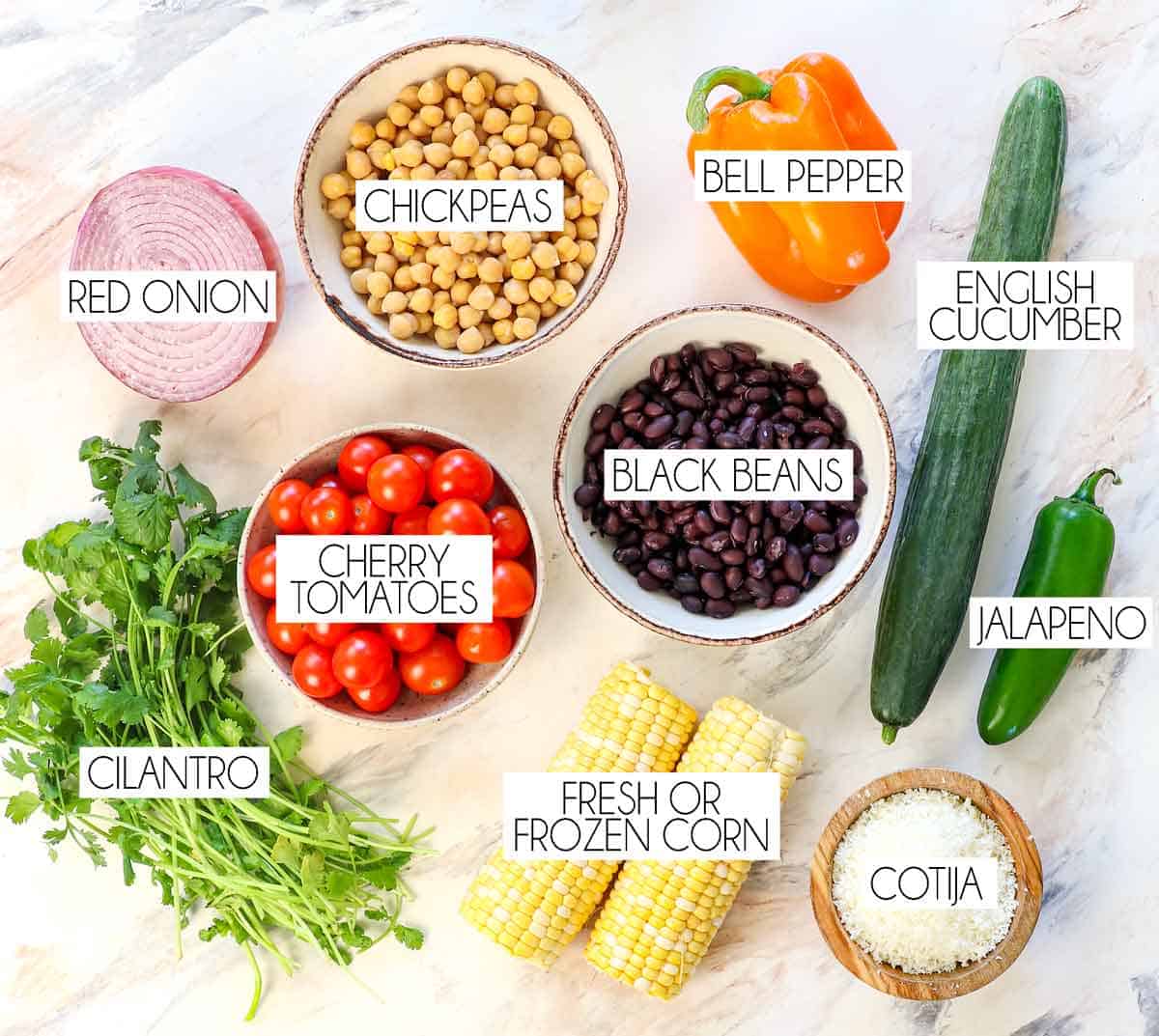 Top view of Dense Bean Salad ingredients: garbanzo beans, black beans, corn, tomatoes, bell pepper, red onion, cucumber, jalapeno, Cotija, cilantro