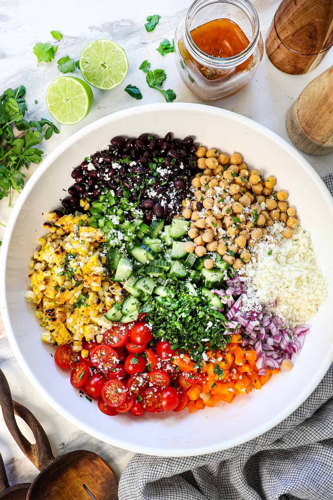 showing how to make dense bean salad recipe by adding the ingredients to a bowl: ichickpeas, black beans, corn, tomatoes, red onion, bell pepper, cotija and cilantro