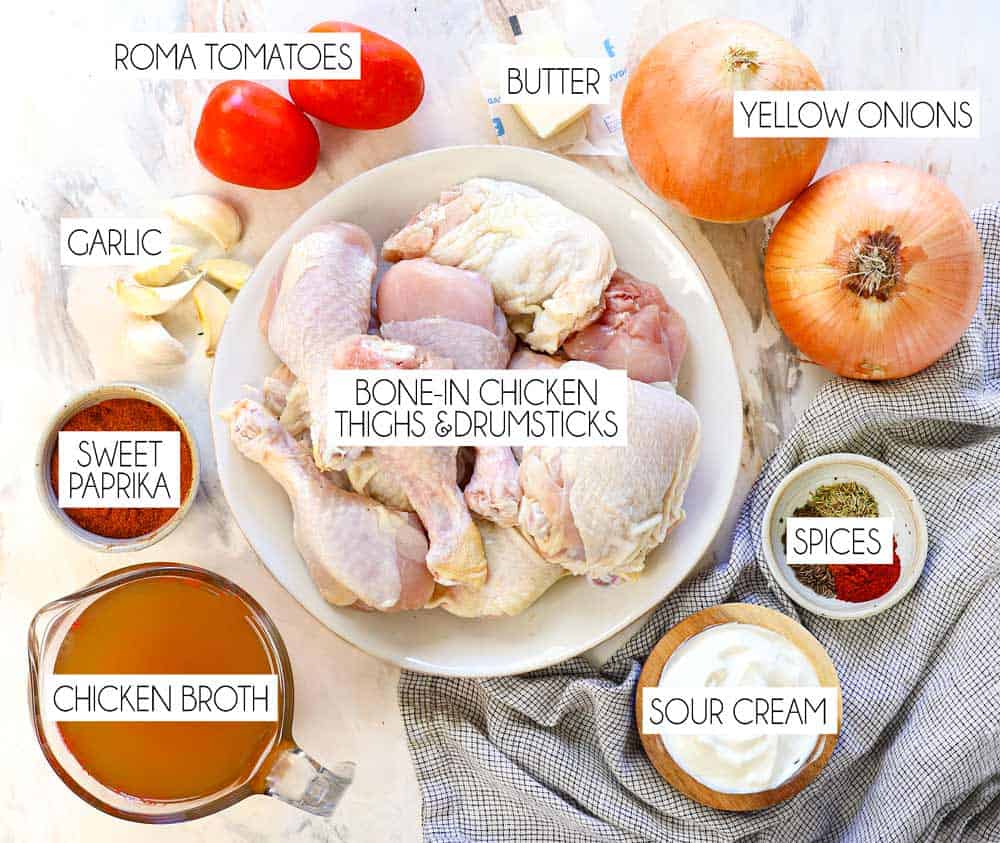 top view of chicken paprikash ingredients: bone-in chicken drumsticks and thighs, Hungarian sweet paprika, yellow onion, garlic, chicken broth, tomatoes, sour cream