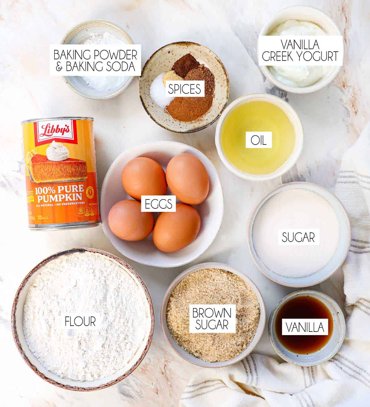 top view of pumpkin cake ingredients: pumpkin puree, eggs, flour, granulated sugar, brown sugar, yogurt, vegetable oil, vanilla extract, baking powder, baking soda, ground cinnamon, ground nutmeg, ground cloves, ground ginger