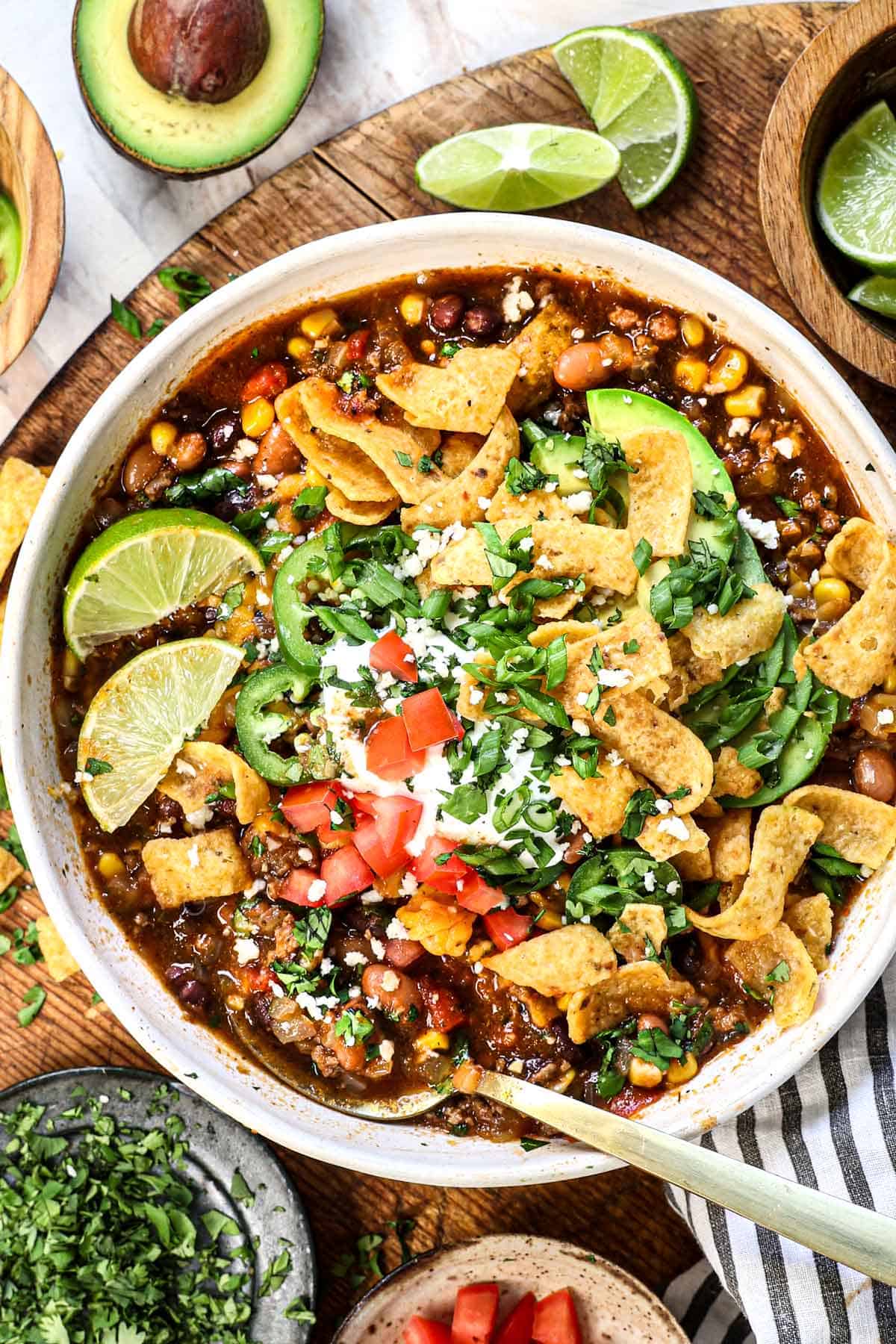 showing how to serve slow cooker taco soup by adding it to a bowl and topping with cheese, sour cream, Fritos chips, tomatoes, cilantro and lime
