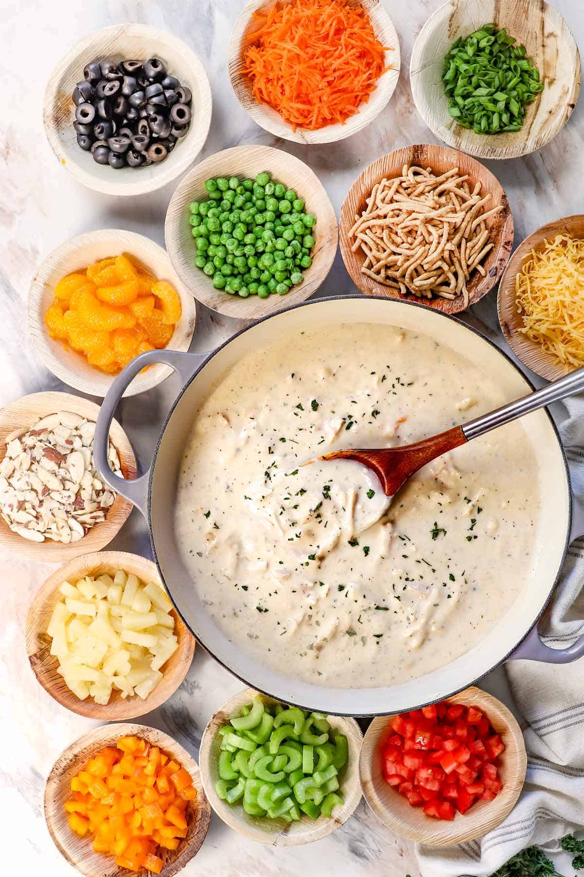 Top view of Hawaiian Haystacks ingredients: creamy chicken gravy, pineapple tidbits, mandarin oranges, frozen peas, chow mein noodles, cheese, sliced almonds, celery, bell peppers. shredded carrots olives, coconut, green onions